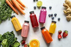 Alt= colorful bottles with smoothies beside carrots ginger leaves and berries